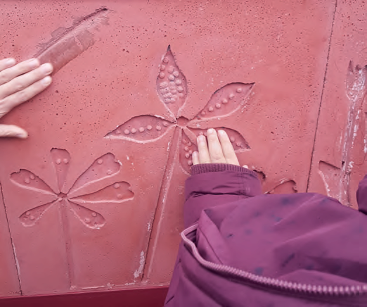 Das Bild zeigt eine verputzte, rötliche Fassade, in die dekorative Blumenmuster in die Oberfläche eingearbeitet bzw. eingeritzt wurden. Eine Kinderhand liegt auf einer dieser Blumenmuster und erfühlt die Struktur. 