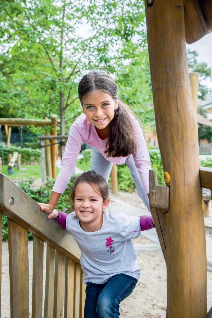 Zwei junge Mädchen sind auf einem Spielsatz. Sie spielen auf einem Klettergerüst. Ein Mädchen geht die Holztreppen des Gerüsts hinaus. Das andere Mädchen steht auf dem Geländer und stützt sich dabei mit den Armen auf dem Geländer ab - es steht über dem anderen Mädchen. Beide lächeln in die Kamera.    